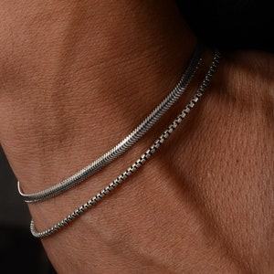 May include: Two silver bracelets on a person's wrist. One bracelet is a thin, snake chain and the other is a thicker, box chain.