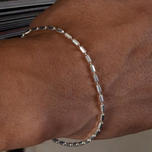 Puede incluir: Un brazalete de cadena de plata con un diseño de cadena de caja. El brazalete está hecho de pequeños eslabones rectangulares.