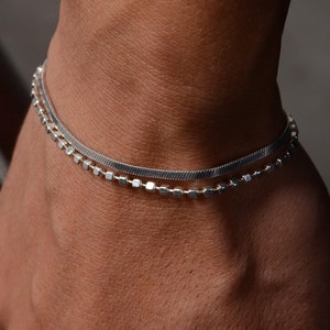 May include: Two silver bracelets on a person's wrist. The top bracelet is a thin, flat, chain-like bracelet. The bottom bracelet is a thicker, square-link chain bracelet.