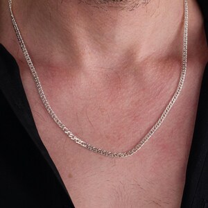 May include: A silver chain necklace with a delicate, intricate design. The chain is worn by a person with a beard and a black shirt.