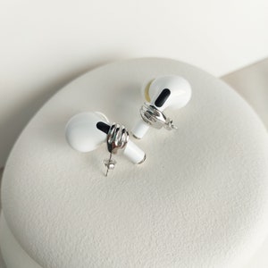May include: A pair of white and silver stud earrings designed to resemble wireless earbuds. Each earring features a silver band and a black detail. The earrings are presented on a white, textured surface.
