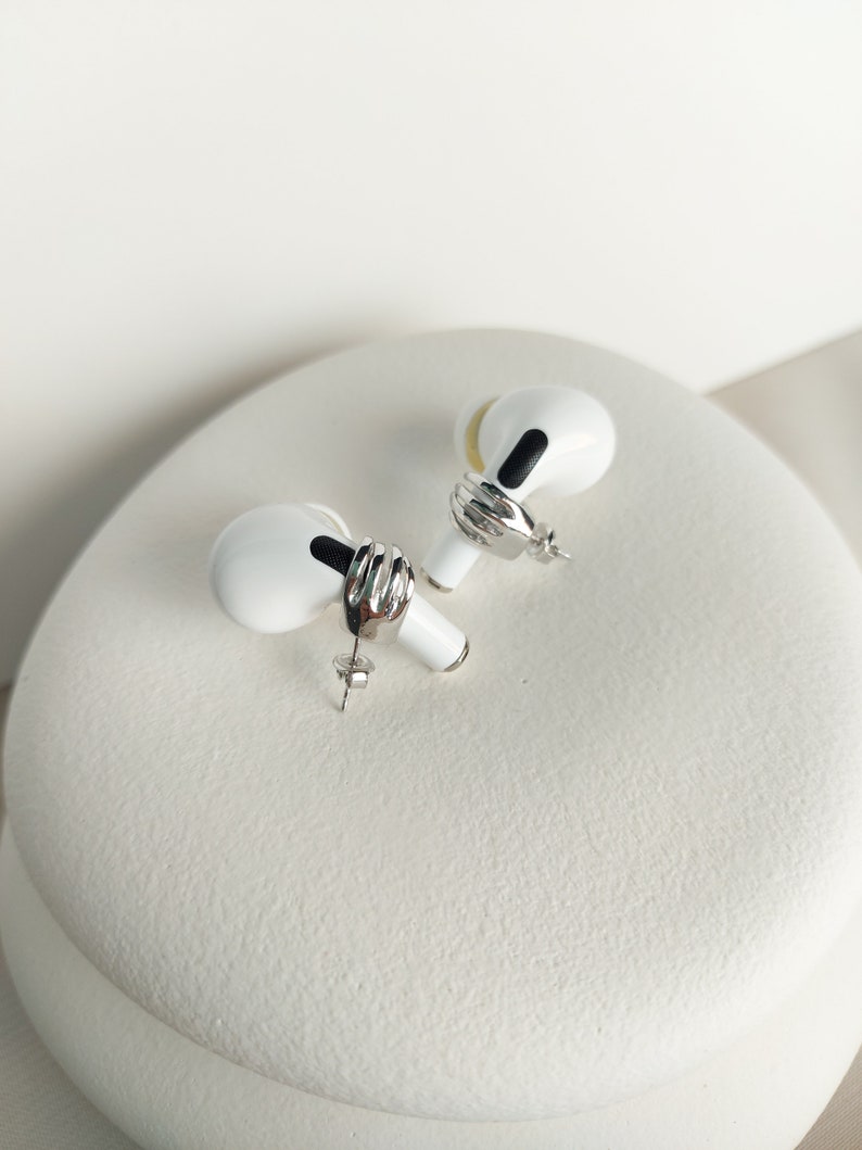 May include: A pair of white and silver stud earrings designed to resemble wireless earbuds. Each earring features a silver band and a black detail. The earrings are presented on a white, textured surface.