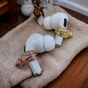 May include: Two white wireless earbuds, each adorned with a decorative hand-shaped ring in gold and rose gold. They are placed on a beige textured surface. The hand-shaped rings add a unique touch to the earbuds.
