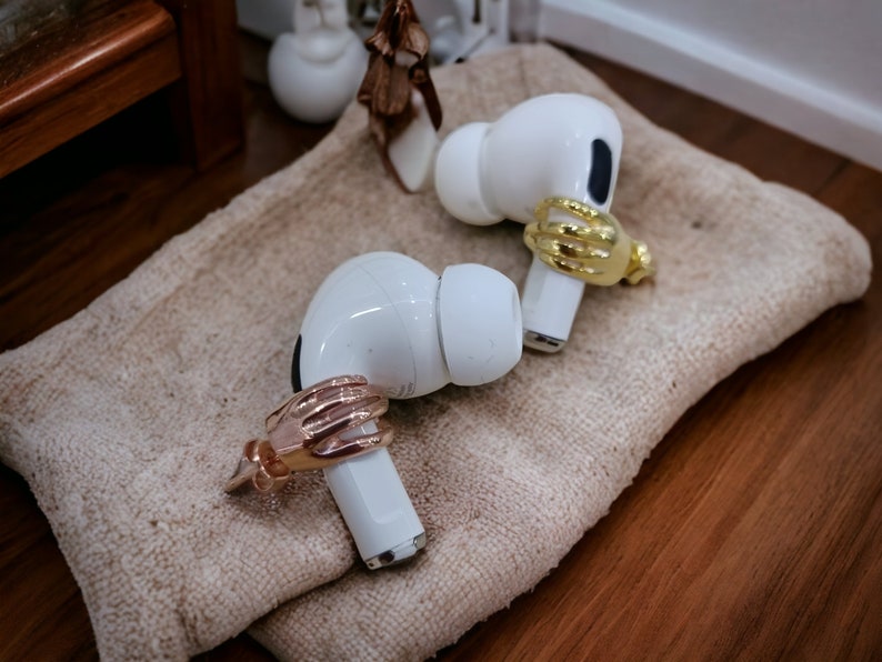 May include: Two white wireless earbuds, each adorned with a decorative hand-shaped ring in gold and rose gold. They are placed on a beige textured surface. The hand-shaped rings add a unique touch to the earbuds.