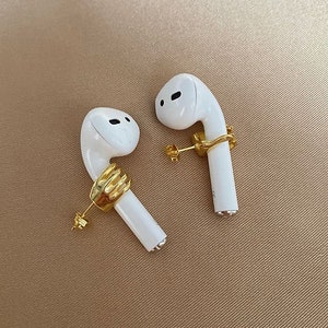 May include: Two white wireless earbuds, each adorned with a gold-coloured hand-shaped earring. The earbuds rest on a tan surface, highlighting the contrast between the white and gold. The earrings provide a unique, artistic touch.