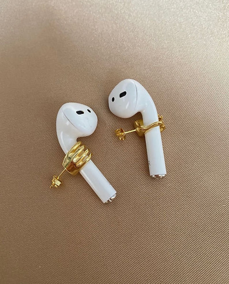 May include: Two white wireless earbuds, each adorned with a gold-coloured hand-shaped earring. The earbuds rest on a tan surface, highlighting the contrast between the white and gold. The earrings provide a unique, artistic touch.