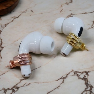 May include: Two white wireless earbuds, each with a hand-shaped accessory. One is gold-coloured, the other rose gold. The earbuds are displayed on a marble surface, highlighting the unique jewellery.