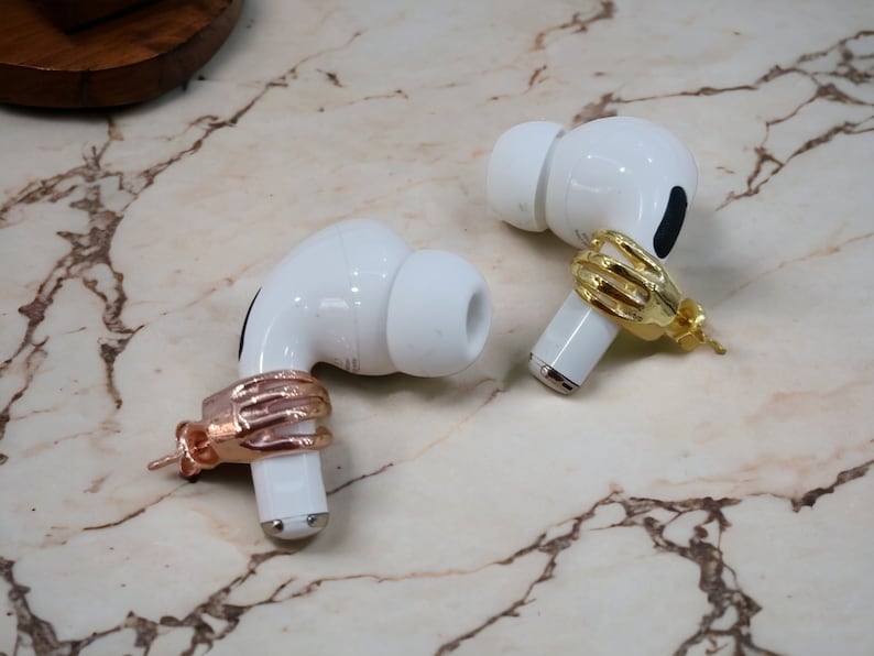 May include: Two white wireless earbuds, each with a hand-shaped accessory. One is gold-coloured, the other rose gold. The earbuds are displayed on a marble surface, highlighting the unique jewellery.