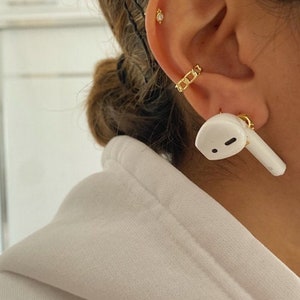 May include: Close-up of a gold chain ear cuff and a small gold stud earring. A white AirPod is in the ear. The person is wearing a white hoodie.