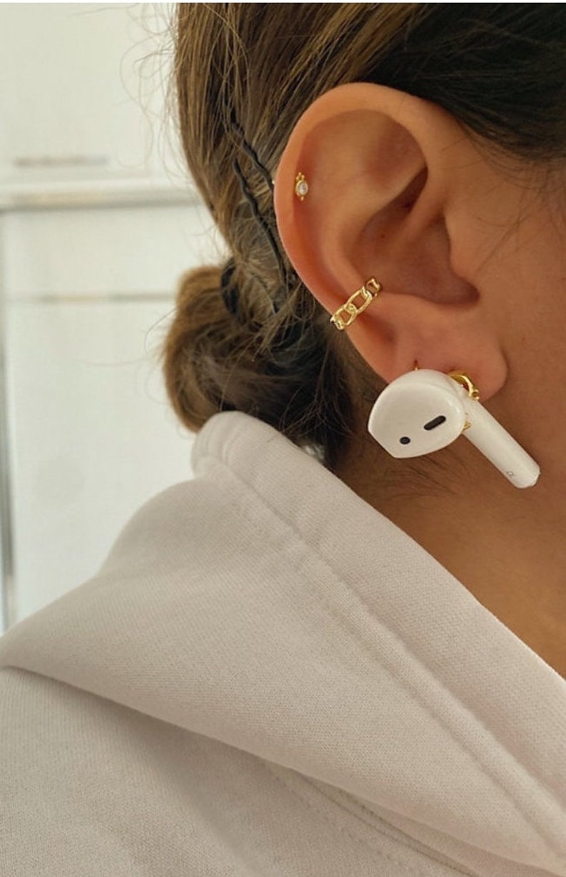 May include: Close-up of a gold chain ear cuff and a small gold stud earring. A white AirPod is in the ear. The person is wearing a white hoodie.