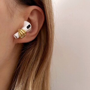 May include: A white earbud with a gold-coloured hand design wrapped around it, worn in an ear. The earbud is set against a backdrop of light brown hair and a light blue top.