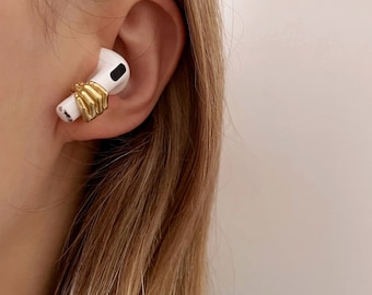 May include: A white earbud with a gold-colored hand design wrapped around it, worn in an ear. The earbud is set against a backdrop of light brown hair and a light blue top.