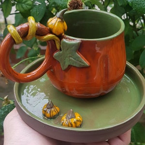 May include: A ceramic pumpkin-shaped coffee mug with a green leaf and a small pumpkin handle. The mug is sitting on a green saucer with two small pumpkins.