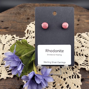 May include: Pink rhodonite stud earrings with sterling silver wire wrapping, displayed on a black card with the text "Rhodonite Emotional Healing Sterling Silver Earrings". The earrings are presented with purple flowers and a lace doily.