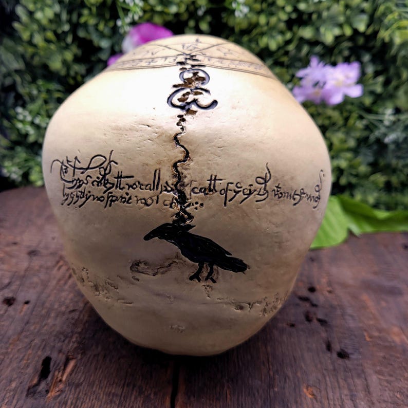 Op de afbeelding: Een cr&egrave;mekleurige schedel met zwarte tekst en een ravenillustratie. De schedel heeft een scheur in het midden en is versierd met ingewikkelde patronen. De achtergrond toont groen gebladerte en paarse bloemen.