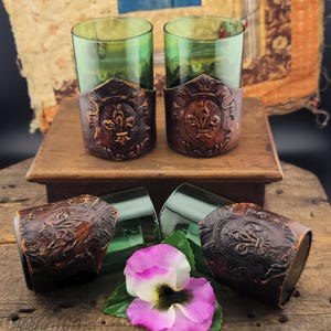 May include: Four green glass tumblers with brown leather wraps featuring a decorative design. The leather wraps have a fleur-de-lis design. The tumblers are sitting on a wooden surface with a purple flower in the foreground.