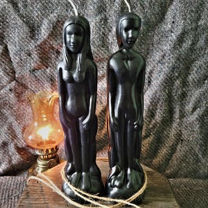May include: Two black wax candles shaped like human figures, one with long hair and one with short hair, standing upright. Each candle features a single white wick at the top and is wrapped with a thin piece of natural twine around the base.