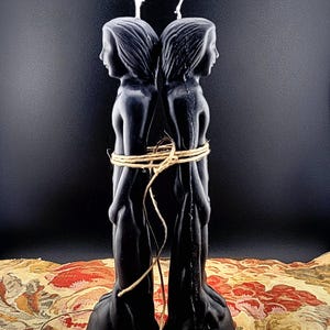 May include: Two black figure candles, each depicting a person, are tied together with twine. The candles are positioned back-to-back, with their profiles facing outward. The background is a dark color, and the candles are placed on a patterned fabric.