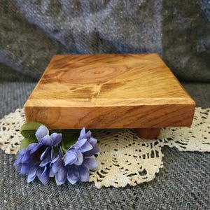 May include: A square wooden riser with a natural light brown wood grain finish and four short cylindrical feet. A cluster of artificial periwinkle blue flowers with green leaves is positioned at the front corner of the wooden platform.