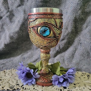 May include: Ornate goblet with a dragon eye design. The chalice features a detailed, textured exterior in gold, bronze, and red hues, with a prominent blue eye. The goblet is topped with a silver rim and rests on a decorative base, with purple flowers.