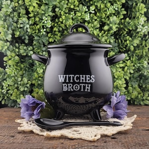May include: A black ceramic cauldron with a lid, handles, and three feet. The words "WITCHES BROTH" are printed on the front in white. A black spoon rests on a cream-colored doily. Purple flowers are on either side of the cauldron.