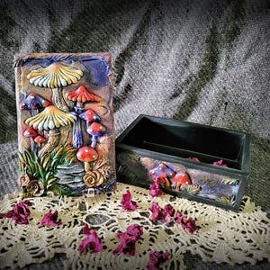 May include: A rectangular black box with a removable lid featuring a relief sculpture of various mushrooms in shades of yellow, red, and purple. The lid displays detailed fungal caps and green foliage, while the box side shows matching mushroom art.