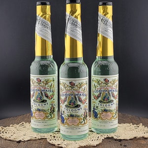 May include: Three glass bottles of Murray & Lanman Florida Water Cologne. The bottles are clear and have a green liquid inside. The bottles have gold foil labels with the text "Murray & Lanman Florida Water Cologne New York" printed on them. The bottles are sitting on a white doily on a wooden surface.