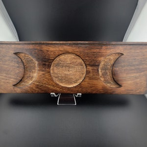 May include: A rectangular wooden box with a dark brown finish. The box features carved crescent moon and circular sun designs. The box is displayed on a clear stand against a black and white background. The box is likely used for storage.