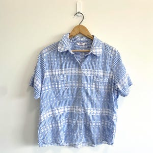 May include: A short-sleeved button-down shirt featuring a light blue and white geometric pattern. The shirt has a standard collar, a front button placket, and two chest pockets. The fabric displays a repeating textured design of stripes.