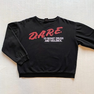 May include: Black sweatshirt with the text "D.A.R.E. TO RESIST DRUGS AND VIOLENCE" in red lettering. The sweatshirt has long sleeves and a crew neck. The text is slightly distressed.