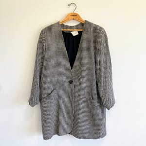 May include: A long-sleeved blazer featuring a black and white houndstooth pattern. The garment has a deep V-neckline, a single central black button closure, and two front pockets. The interior is lined with a solid black fabric.