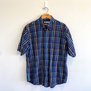 May include: A short-sleeved button-down shirt featuring a dark blue base with a grid pattern of light blue, bright blue, and tan vertical and horizontal lines. The shirt has a pointed collar, a front button placket, and is displayed on a wooden hanger.