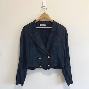 Puede incluir: Una chaqueta corta de color azul marino oscuro con un patrón jacquard floral tonal, solapa de muesca, mangas largas y un cierre frontal de doble botonadura con cuatro botones redondos de color claro. La prenda cuelga de una percha de madera.