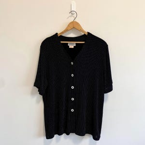 May include: A black short-sleeved button-down shirt featuring a fine white polka dot pattern. The garment has a V-neckline, five white buttons down the centre front, and a label at the inner collar that reads SPORT. The shirt is displayed on a hanger.