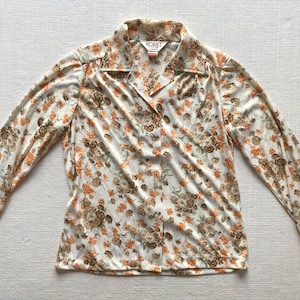 May include: A long-sleeved button-down shirt in an off-white colour featuring a repeating floral pattern in shades of orange and brown. The shirt has a pointed collar and a label at the neckline that reads KORET CANADA.