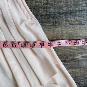 May include: A light pink garment with soft vertical pleats and a visible pocket opening. A pink measuring tape is positioned horizontally across the fabric, showing a measurement extending from 16 inches to 25 inches across the width of the item.