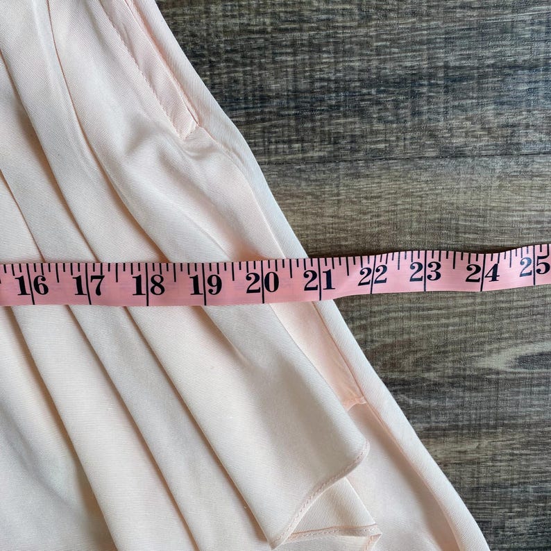 May include: A light pink garment with soft vertical pleats and a visible pocket opening. A pink measuring tape is positioned horizontally across the fabric, showing a measurement extending from 16 inches to 25 inches across the width of the item.