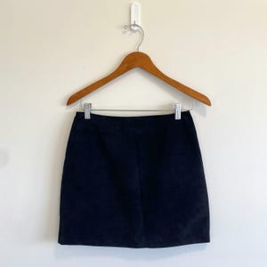Peut inclure: Une mini-jupe en daim bleu marine foncé uni avec un ourlet droit. La jupe est suspendue à un cintre en bois doté d'un crochet en métal argenté et de deux pinces en métal argenté qui maintiennent fermement le bord supérieur du tissu.