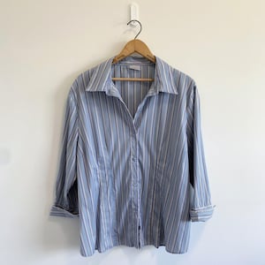 May include: A long-sleeve button-down shirt featuring a vertical striped pattern in shades of light blue, dark blue, white, and thin tan lines. The shirt has a classic collar, a front button placket, and cuffs at the end of the sleeves.