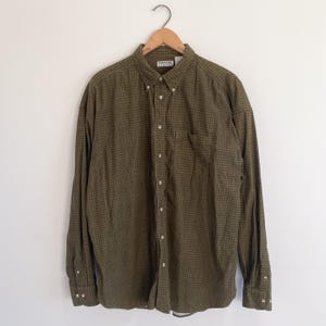 Könnte beinhalten: Ein langärmeliges Button-Down-Hemd mit einem olivgrünen und dunkelbraunen Mikro-Karomuster. Das Hemd verfügt über einen Button-Down-Kragen, eine einzelne Brusttasche und helle Knöpfe. Ein weißes Etikett mit dem Text Northern Elements ist sichtbar.