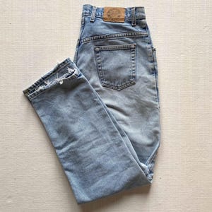 May include: Light blue denim jeans with a faded wash and a distressed hem. The jeans have a classic five-pocket design and a leather patch on the back waistband. The jeans are folded on a light-coloured surface.