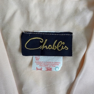 May include: A light peach-colored garment featuring a dark navy blue rectangular clothing label with the brand name Chablis embroidered in gold script. Below the main label is a white tag with red text reading Made in Canada and Exclusive of Trim.