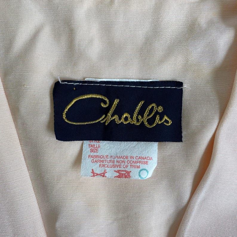 May include: A light peach-colored garment featuring a dark navy blue rectangular clothing label with the brand name Chablis embroidered in gold script. Below the main label is a white tag with red text reading Made in Canada and Exclusive of Trim.