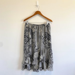 May include: A knee-length skirt featuring a black and white floral pattern with a geometric background print. The skirt has an elasticized waistband and a tiered, ruffled hemline, hanging from a wooden hanger against a plain white wall.