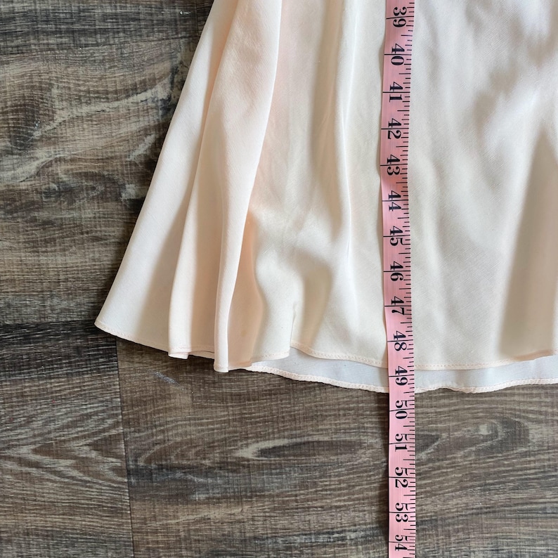 May include: A pale peach-colored skirt with a flared, flowing silhouette. A pink measuring tape is placed vertically along the fabric, showing a length measurement extending from 39 inches down to 54 inches along the hemline of the garment.