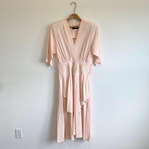 May include: A pale pink midi dress featuring a V-neckline, elbow-length sleeves, and a fitted waist with delicate white floral embroidery. The skirt has a layered, ruffled design that falls below the knees, creating a soft, flowing silhouette.