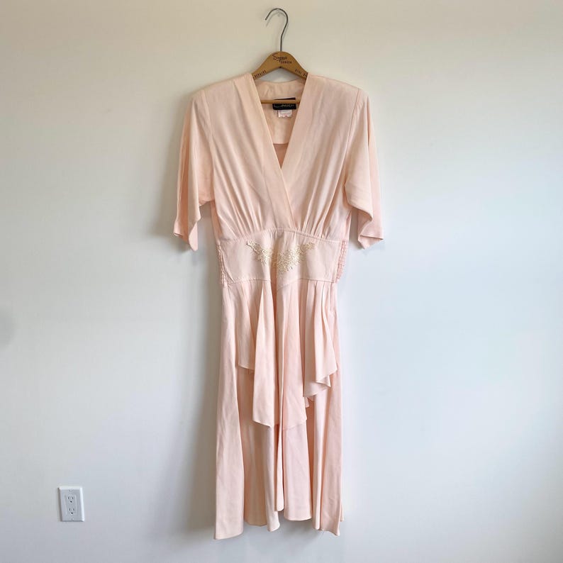 May include: A pale pink midi dress featuring a V-neckline, elbow-length sleeves, and a fitted waist with delicate white floral embroidery. The skirt has a layered, ruffled design that falls below the knees, creating a soft, flowing silhouette.