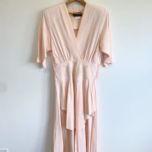 May include: A pale peach-colored dress with a V-neckline, elbow-length sleeves, and a fitted waist featuring a central cream-colored floral embroidery detail. The skirt has a layered, asymmetrical hemline with soft pleats and gathers at the waist.