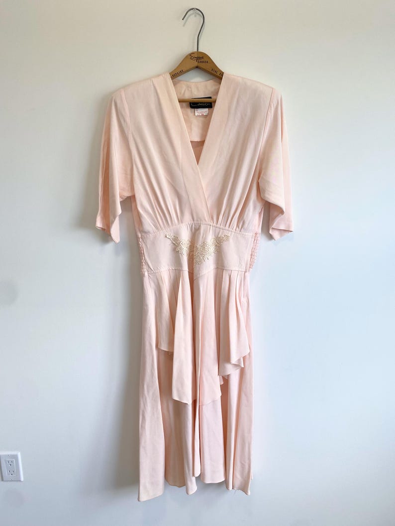 May include: A pale peach-colored dress with a V-neckline, elbow-length sleeves, and a fitted waist featuring a central cream-colored floral embroidery detail. The skirt has a layered, asymmetrical hemline with soft pleats and gathers at the waist.