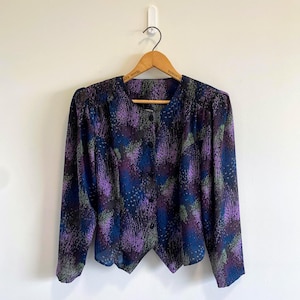 May include: A long-sleeved button-down blouse featuring a dark base with an abstract pattern of purple, blue, and green speckles. The garment has a round neckline, gathered shoulders, and a pointed hemline, hanging on a wooden hanger.